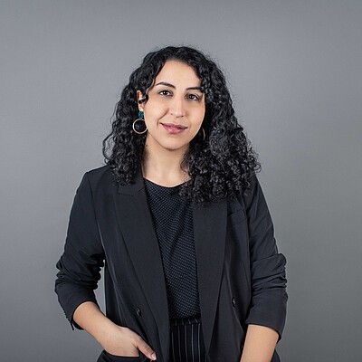 Emira Abidi Weibliche Person, lächelnd, dunkles Oberteil, dunkle lockige Haare