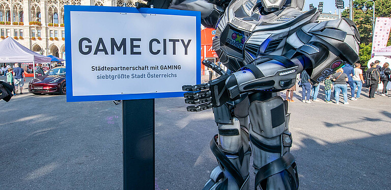 Person im Roboter Kostüm zeigt auf Schild mit Text "Game City"