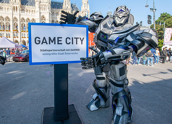 Person im Roboter Kostüm zeigt auf Schild mit Text "Game City"