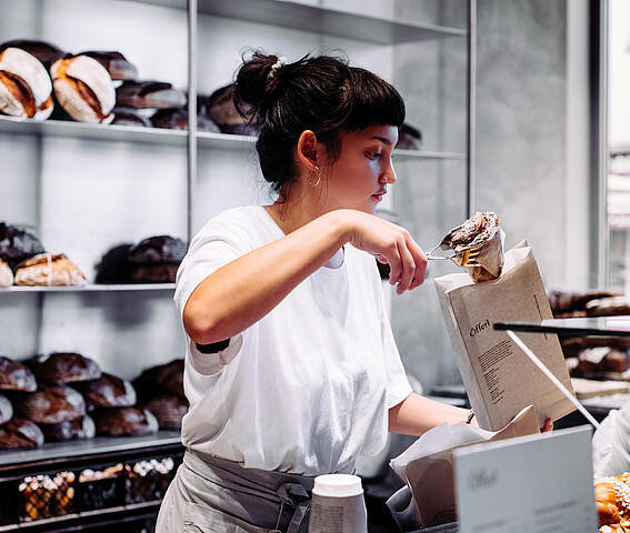 Graetzel beleben: Förderungen, Beratungen, Netzwerke für Unternehmen/Nahversorger*innen in Wien. Eine Frau in einer Bäckerei steckt ein Stück Gebäck in ein Verkaufssackerl.