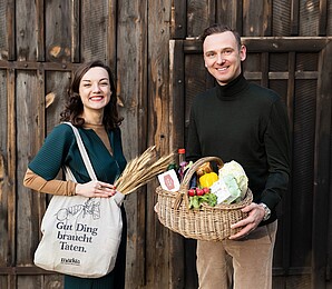 markta: Eine Frau und ein Mann stehen mit einem Getreidesack und einem Obstkorb vor einer Scheunentüre aus Holz. 